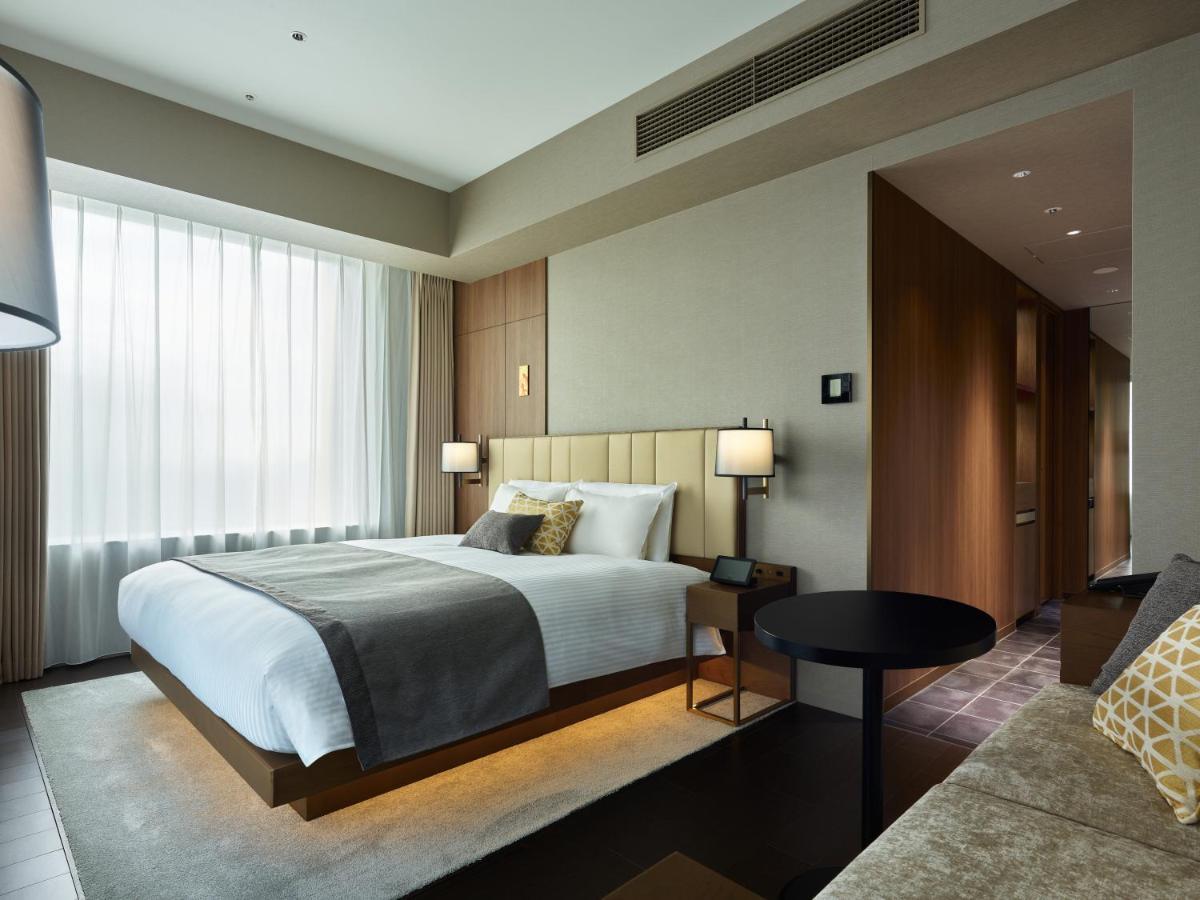 Mitsui Garden Hotel Roppongi Tokyo Premier Pokój zdjęcie The photo shows a modern hotel room. It features a large bed with a light-colored duvet and decorative pillows. There are bedside tables with lamps on either side of the bed. The room has large windows dressed with sheer curtains, allowing natural li