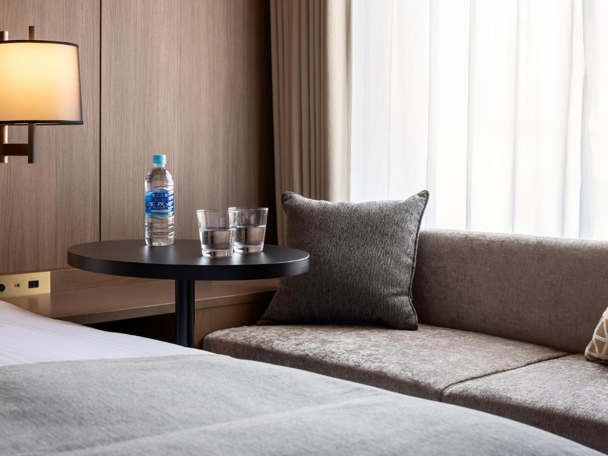 Mitsui Garden Hotel Roppongi Tokyo Premier Zewnętrze zdjęcie The photo depicts a cozy hotel room setting. On the left, there is a bedside table with a lamp. Against the wall, there is a comfortable-looking couch with a decorative pillow. In front of the couch, there's a small round table that holds a bottle of