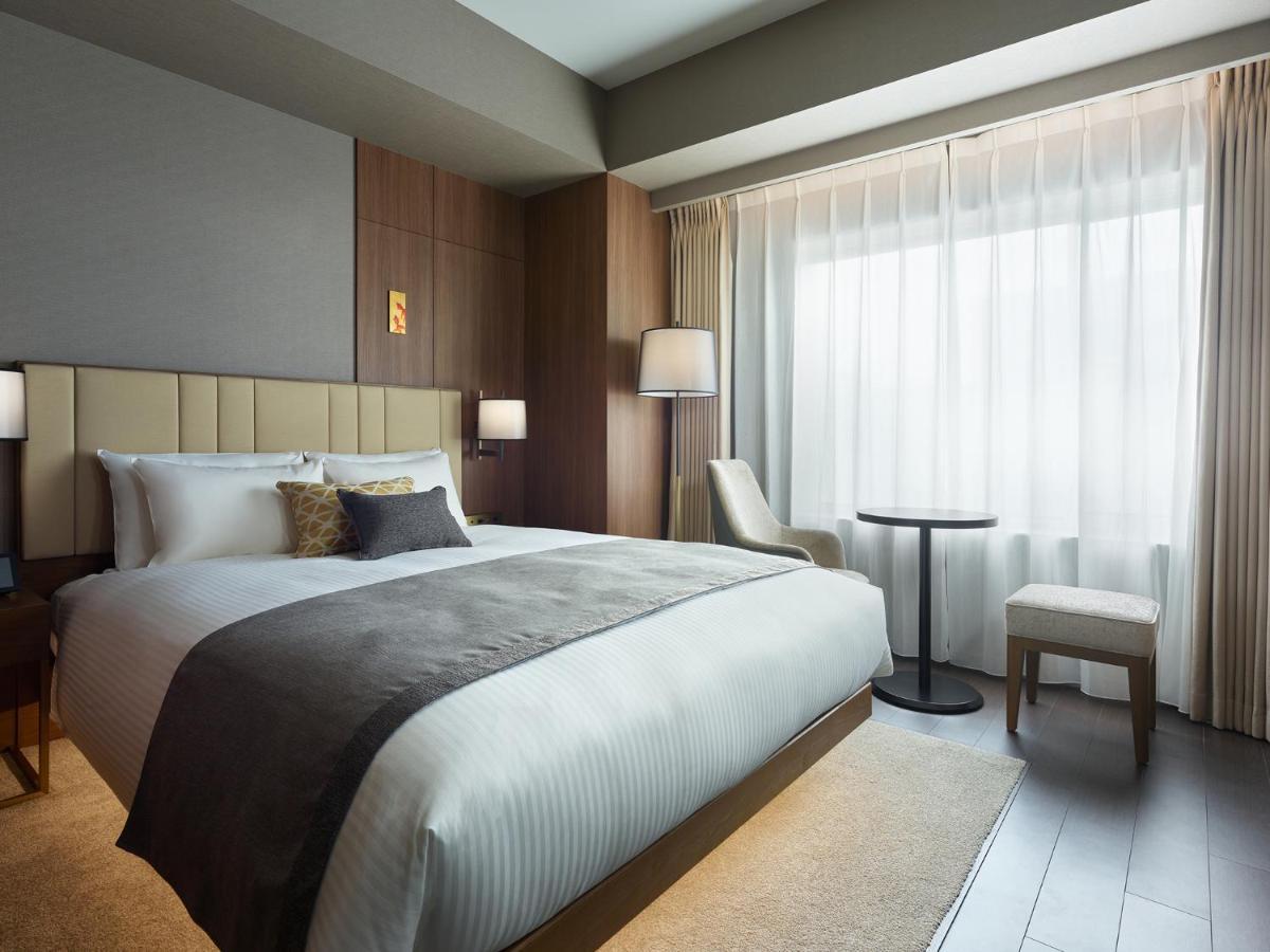 Mitsui Garden Hotel Roppongi Tokyo Premier Zewnętrze zdjęcie The photo shows a modern hotel room featuring a comfortable bed with white linens and a decorative throw pillow. The room has a neutral color palette with wooden accents on the walls. There are two bedside lamps and a chair positioned next to a small