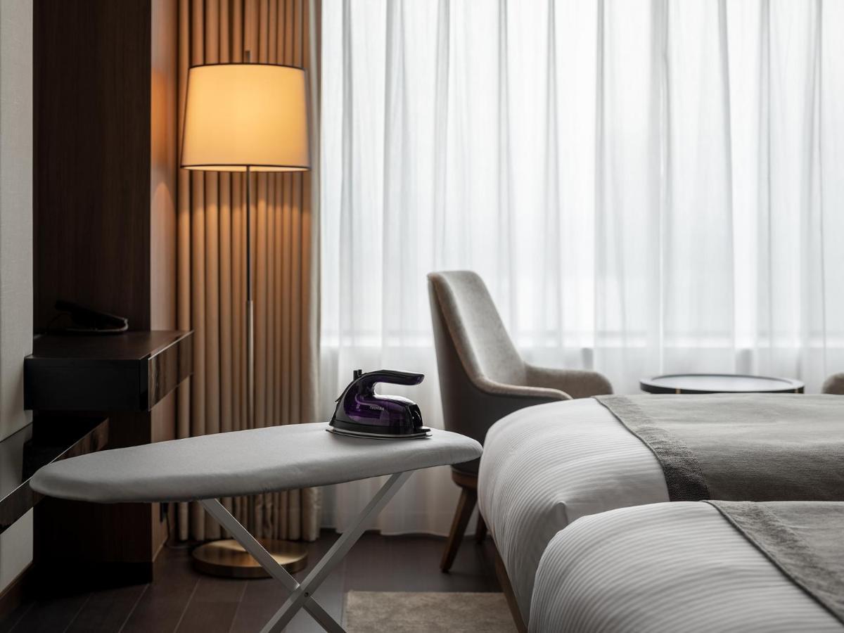Mitsui Garden Hotel Roppongi Tokyo Premier Zewnętrze zdjęcie The photo shows a modern hotel room featuring two beds with white bedding. There is a light-colored armchair next to a small side table. On the table, there is an iron, and beside it, a lamp with a warm glow is illuminating the space. The large windo