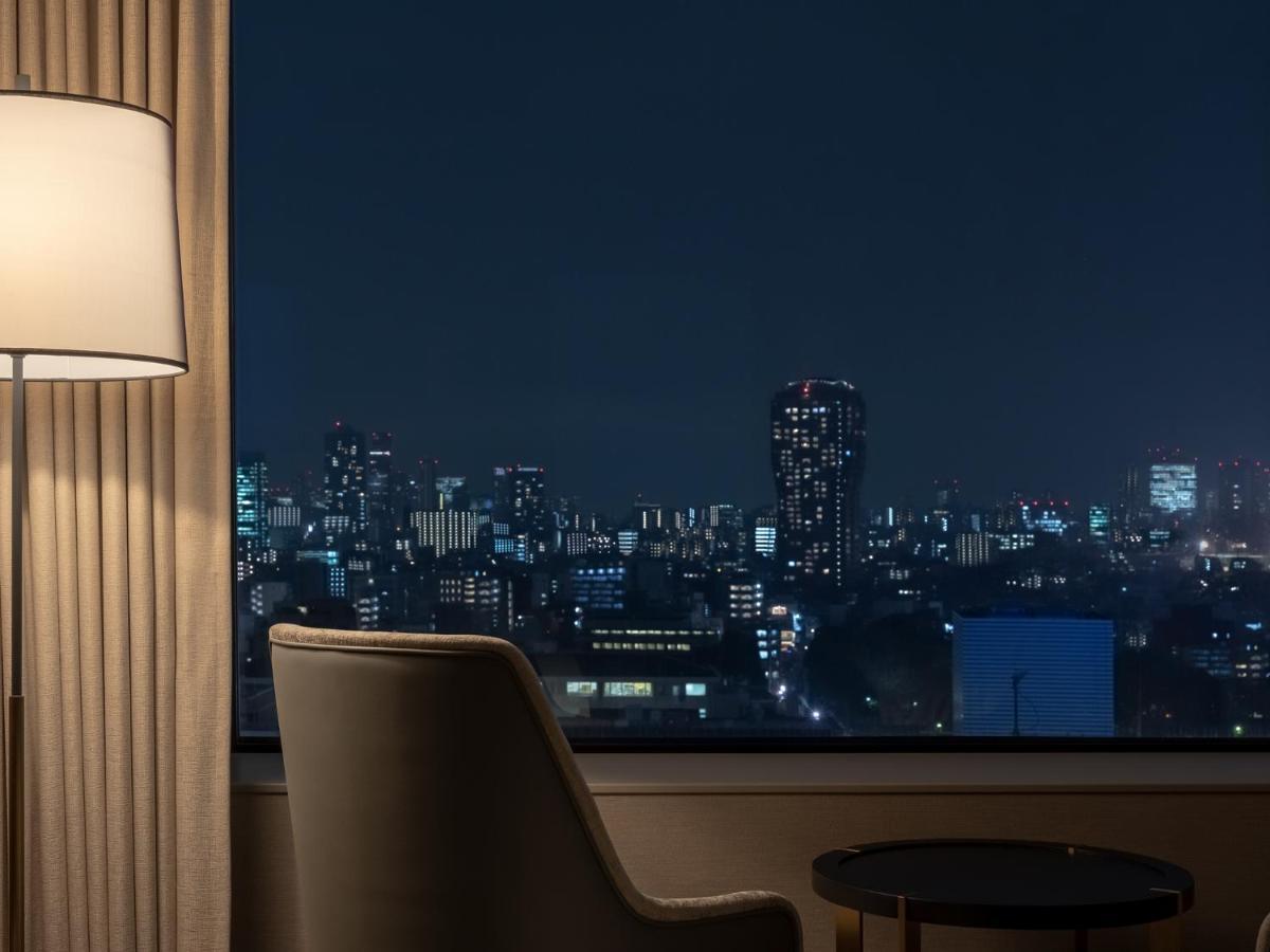 Mitsui Garden Hotel Roppongi Tokyo Premier Zewnętrze zdjęcie The photo depicts a cozy hotel room setting at night. In the foreground, there's a stylish armchair next to a small round table. A standing lamp provides soft lighting, creating a warm ambiance. In the background, large windows reveal a city skyline 