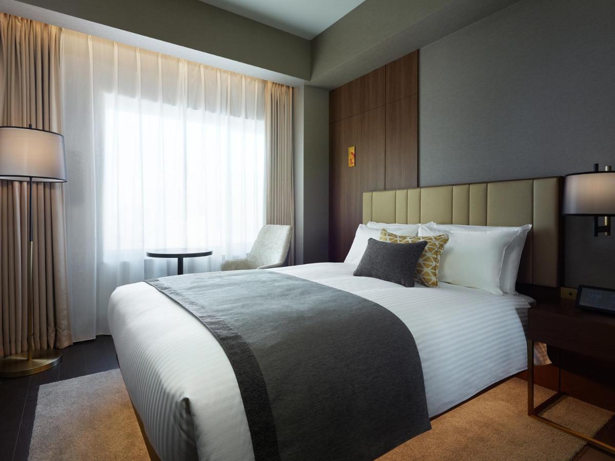 Mitsui Garden Hotel Roppongi Tokyo Premier Zewnętrze zdjęcie The photo shows a modern hotel room featuring a neatly made bed with white linens and a decorative gray throw blanket. To the side, there is a small round table and a comfortable chair, creating a cozy seating area. The room is well-lit with natural 