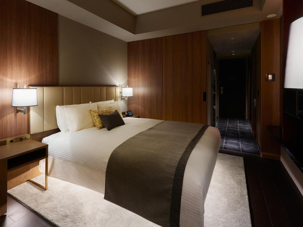 Mitsui Garden Hotel Roppongi Tokyo Premier Zewnętrze zdjęcie The image shows a modern hotel room with a warm and elegant design. In the center, there is a neatly made bed with white linens and a dark gray throw blanket. Flanking the bed are two bedside lamps providing soft lighting. The walls have a rich wood 