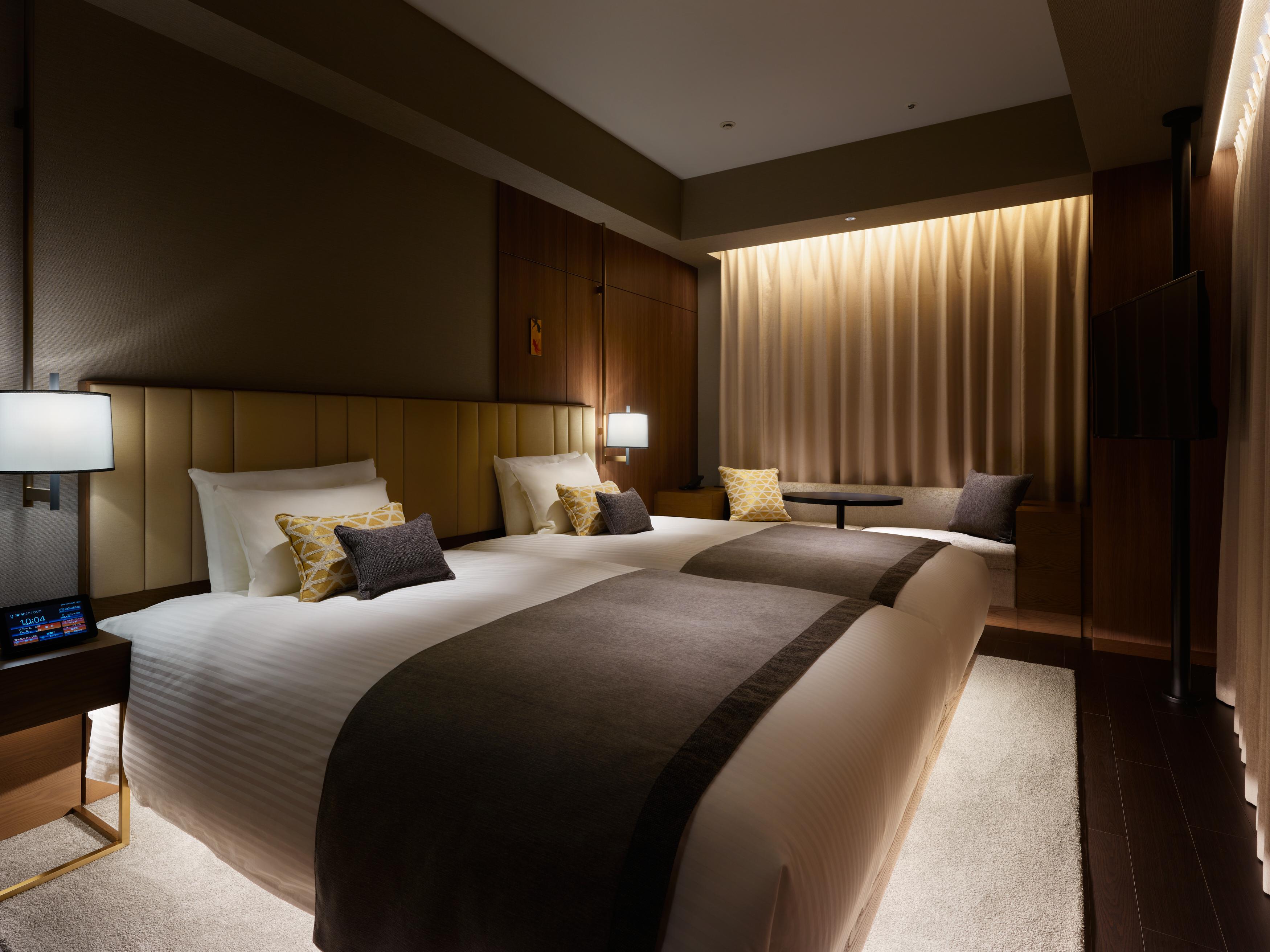 Mitsui Garden Hotel Roppongi Tokyo Premier Zewnętrze zdjęcie The photo shows a modern hotel room featuring two beds with plush white linens and decorative pillows. The room has a warm and inviting ambiance, highlighted by soft lighting from lamps and the backlit curtains. There is also a small table with chair