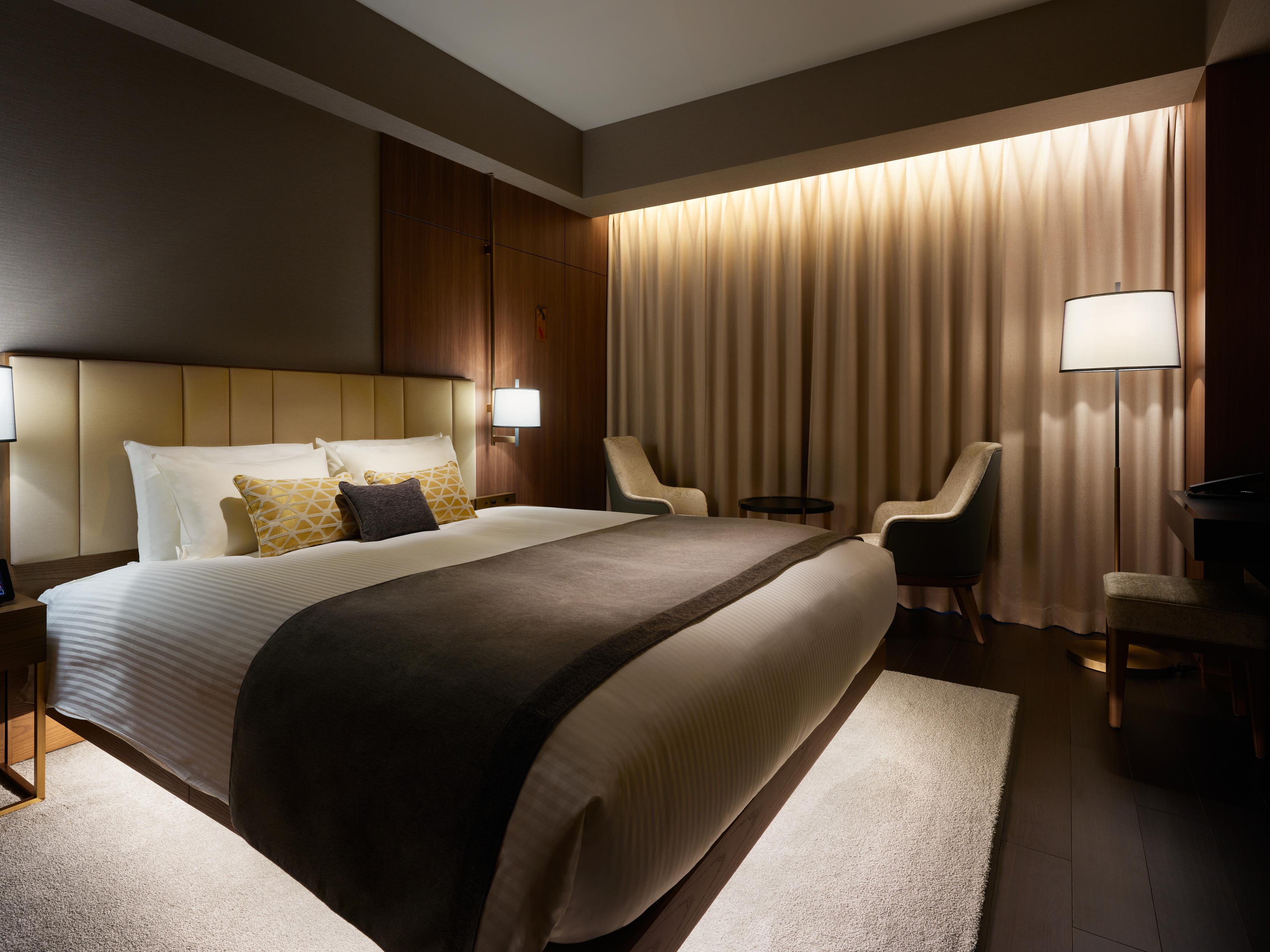 Mitsui Garden Hotel Roppongi Tokyo Premier Zewnętrze zdjęcie The photo depicts a modern hotel bedroom. It features a large bed with white linens and a dark gray throw blanket, accented by decorative pillows. The room has a warm ambiance created by soft lighting, including two bedside lamps and a lighted area b