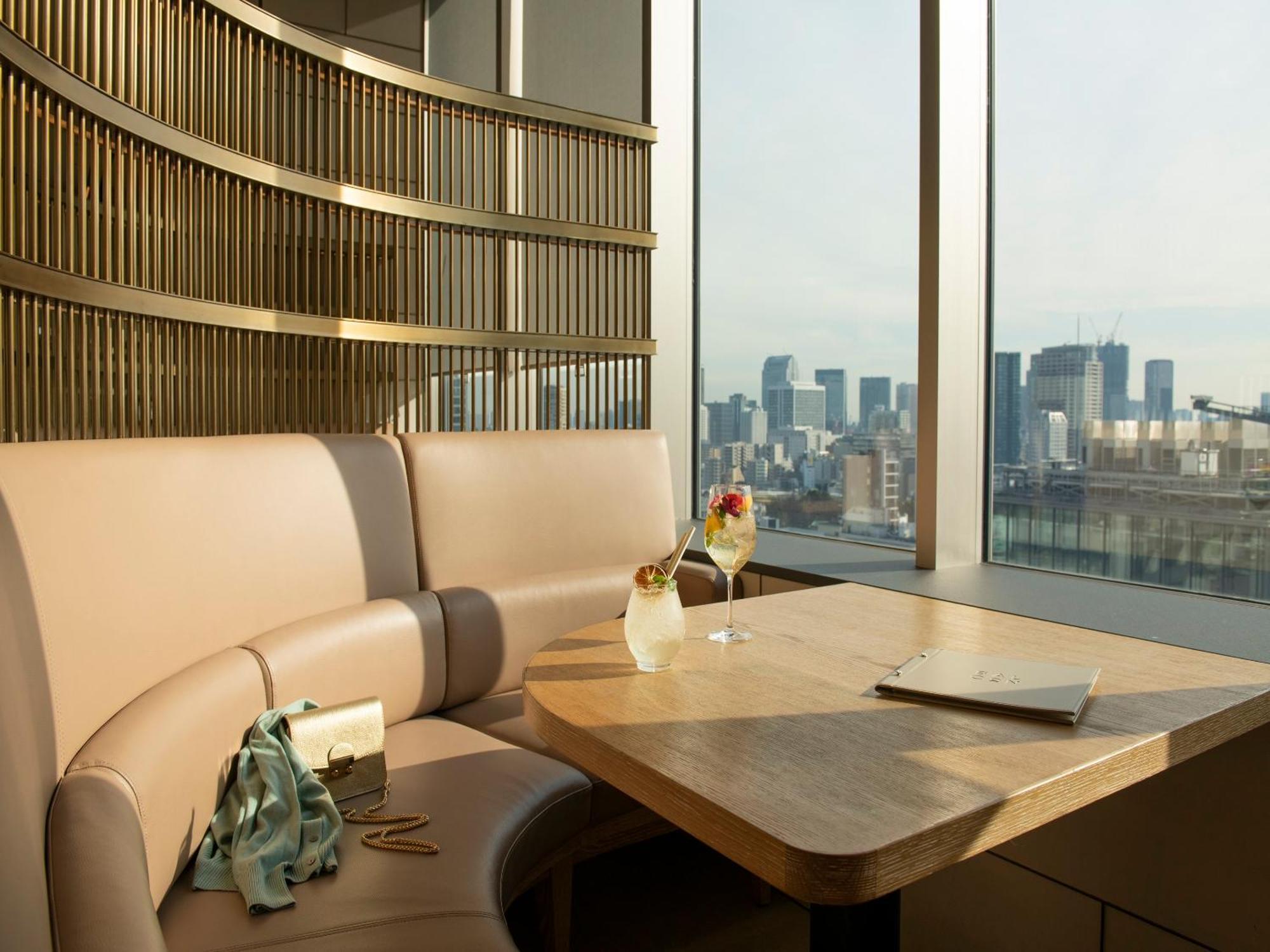 Mitsui Garden Hotel Roppongi Tokyo Premier Zewnętrze zdjęcie The photo depicts a cozy, modern seating area with a curved booth that features soft, pale upholstery. A wooden table is positioned in front of the booth, and on the table, there are two elegant drinks garnished with fruit. The setting has large wind