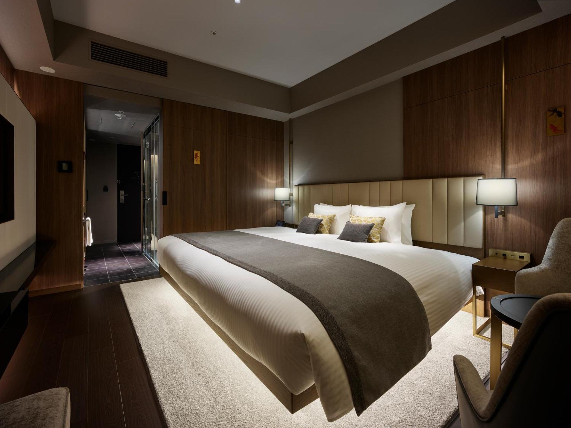Mitsui Garden Hotel Roppongi Tokyo Premier Zewnętrze zdjęcie The photo shows a modern hotel room featuring a large bed with a neutral-colored duvet and a dark throw blanket. The headboard is upholstered, and there are two bedside lamps on either side of the bed. The walls have a warm wood paneling, contributin