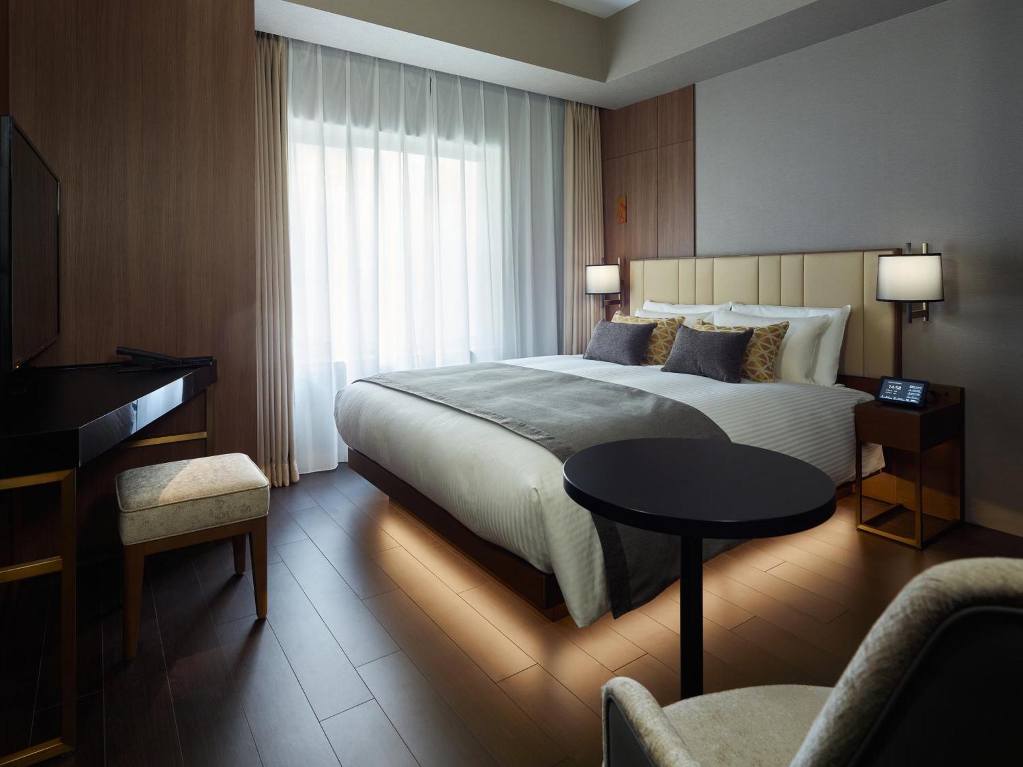 Mitsui Garden Hotel Roppongi Tokyo Premier Zewnętrze zdjęcie The photo shows a modern hotel room featuring a large bed with decorative pillows. There are large windows with sheer curtains allowing natural light to filter in. To one side, there is a sleek desk with a chair and a small round table in the center 