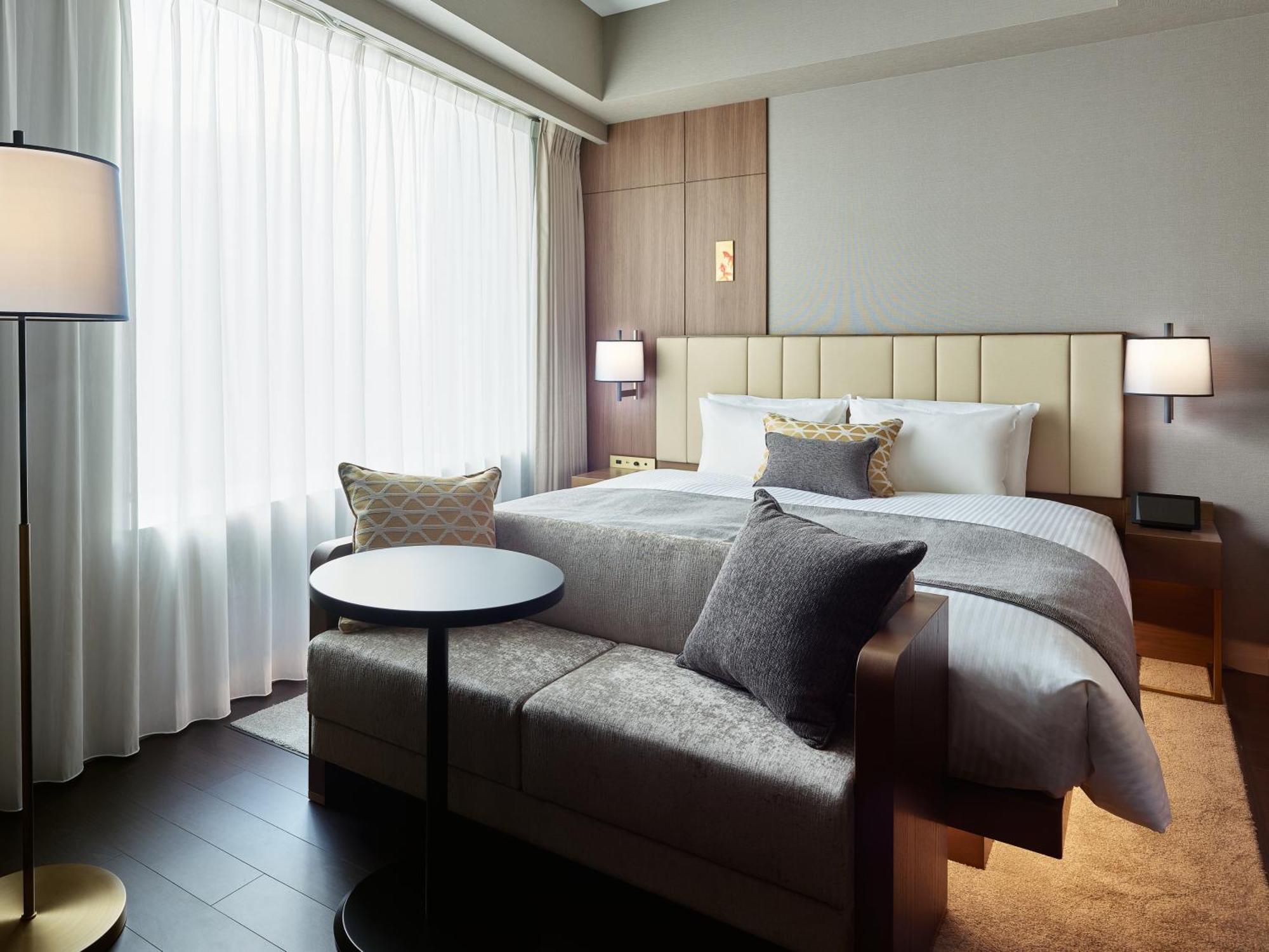 Mitsui Garden Hotel Roppongi Tokyo Premier Zewnętrze zdjęcie The photo shows a modern hotel room that features a large bed with a padded headboard and soft bedding. There's a small, round table accompanied by a couch or seating area, adorned with decorative pillows. To the left, a floor lamp provides additiona