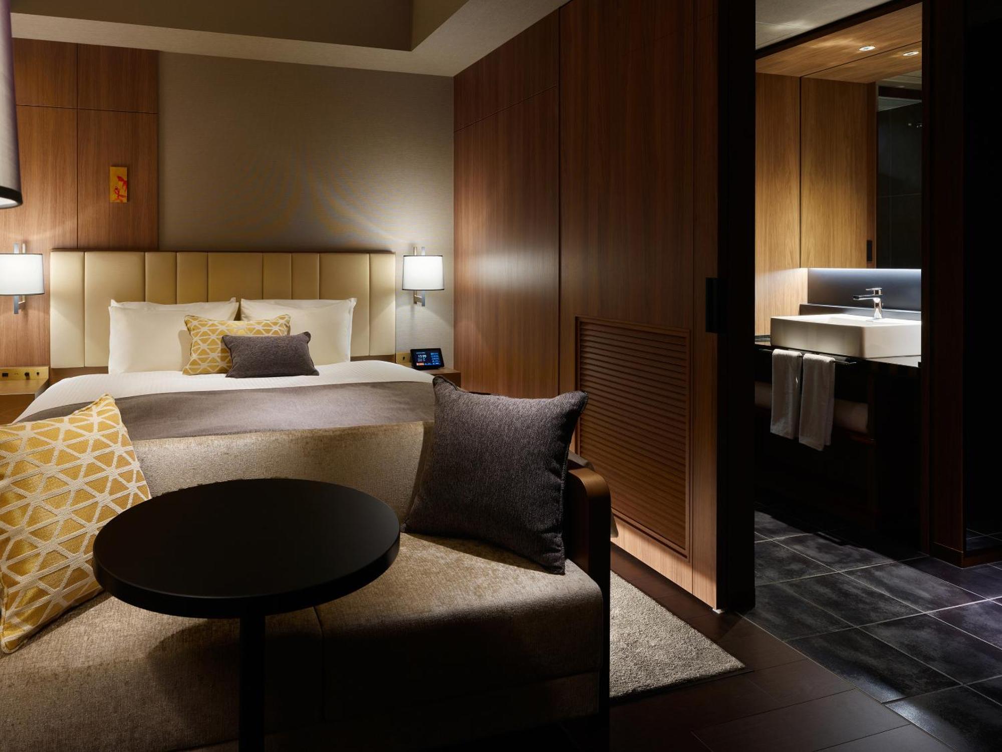 Mitsui Garden Hotel Roppongi Tokyo Premier Zewnętrze zdjęcie The photo depicts a modern hotel room featuring a bed with a stylish headboard and decorative pillows. On one side of the bed, there are bedside lamps providing soft lighting. There is a comfortable seating area with a sofa adorned with pillows, and 