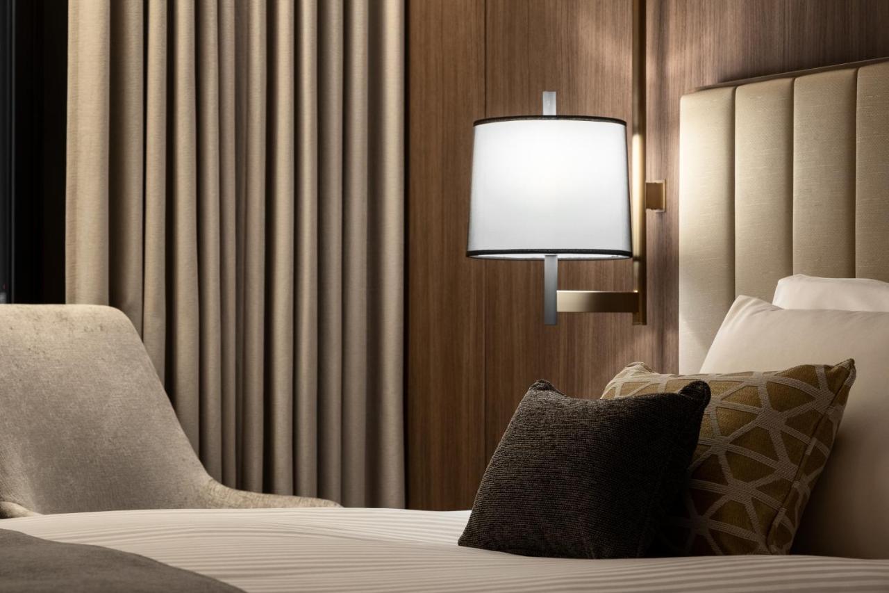 Mitsui Garden Hotel Roppongi Tokyo Premier Zewnętrze zdjęcie The photo shows a cozy hotel bedroom setting. There is a neatly made bed with white linens and a few decorative pillows, including a dark one and another with a geometric pattern. To the side, there is a soft armchair and a modern wall lamp mounted a